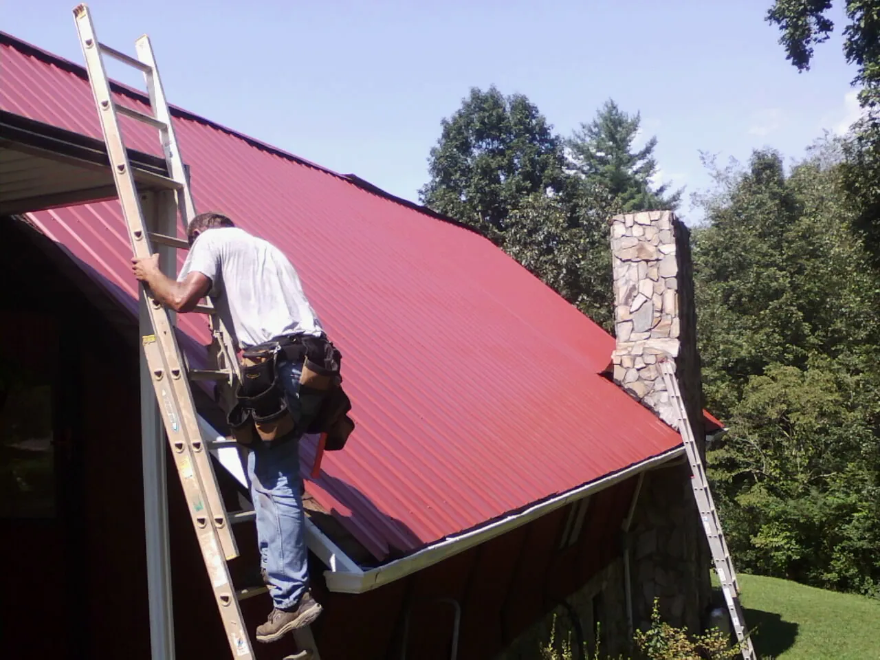 Roofer on a ladder beside a red metal roof and stone chimney — Soffit & Fascia Repair by Roofing Brothers in Cullowhee